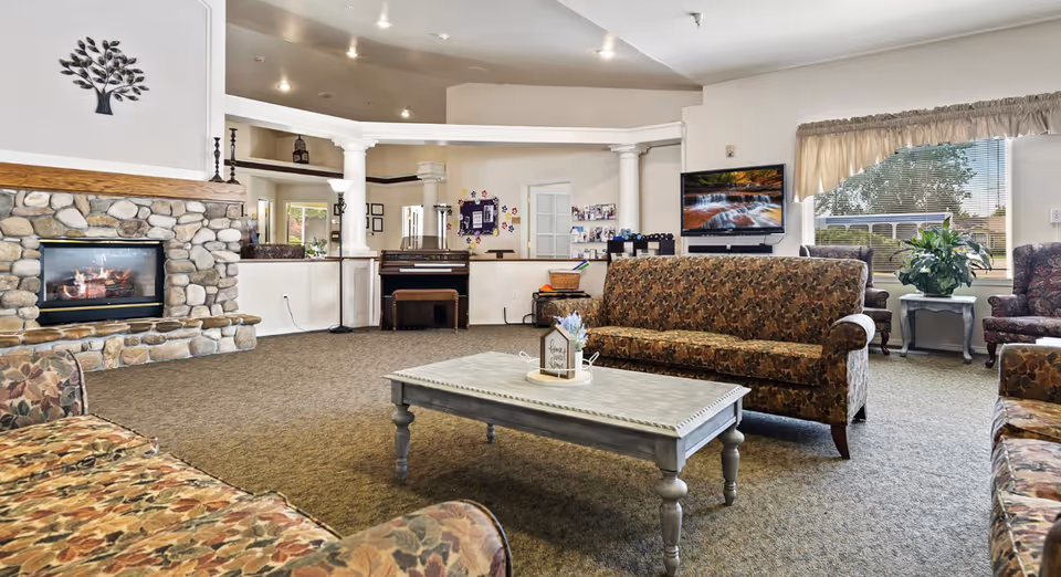 A spacious living room in Autumn Wind Assisted Living featuring floral patterned sofas arranged around a light gray coffee table. There is a stone fireplace with a fire burning on the left side, a piano against the far wall, and a flat-screen TV mounted on the wall showing a nature scene. Large windows with beige curtains let in natural light, and there are decorative plants and wall art enhancing the cozy atmosphere.