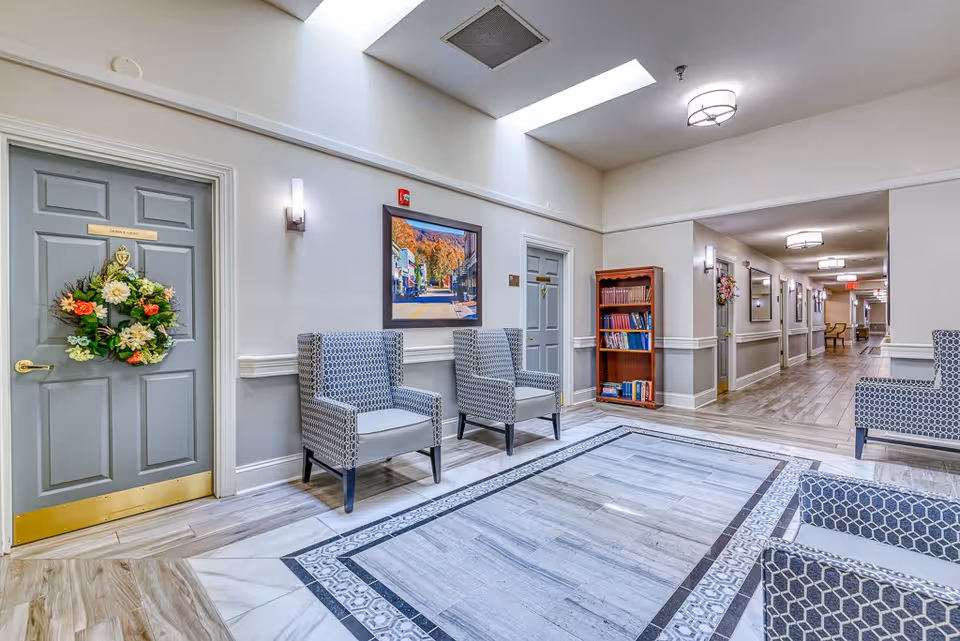A bright and clean senior living facility hallway with patterned armchairs arranged around a decorative tiled floor area. There are two gray doors, one adorned with a floral wreath and a sign that reads 'Donna Gale'. A wooden bookshelf filled with books is placed against the wall. The hallway extends into the distance with more chairs and framed pictures on the walls under ceiling lights.