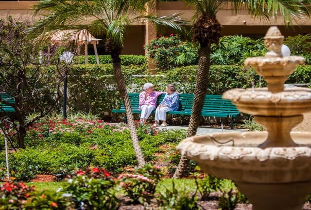 Two elderly women sitting and talking on a green bench in a lush garden area with palm trees, flowering plants, and a decorative stone fountain in the foreground.