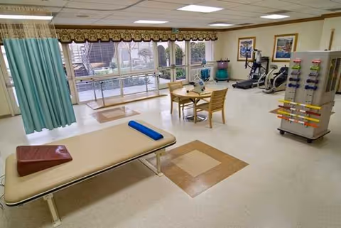 Rehabilitation therapy room with a treatment table, exercise equipment, and seating near windows.