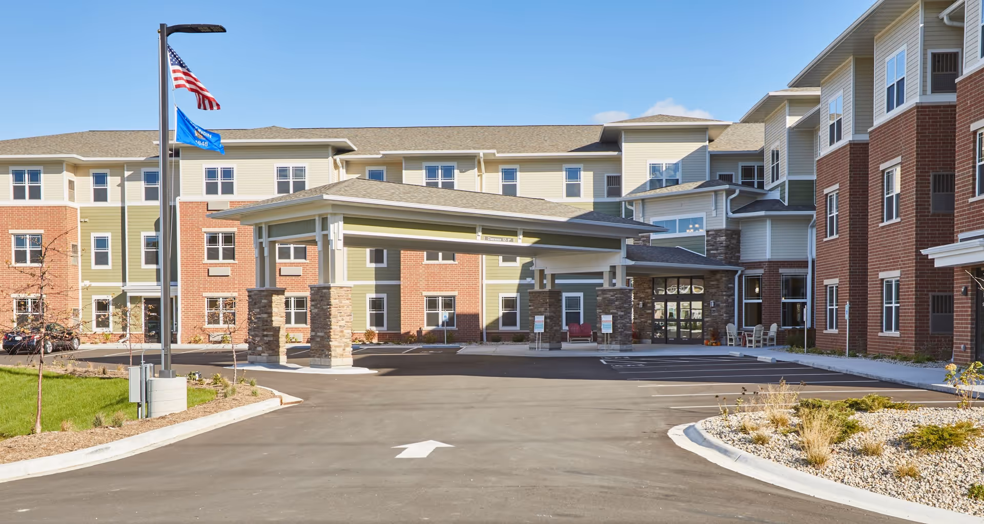 Front exterior view of StoryPoint Mequon senior living facility with a covered entrance, American and Wisconsin state flags on a flagpole, multiple windows, and a parking area with landscaping.