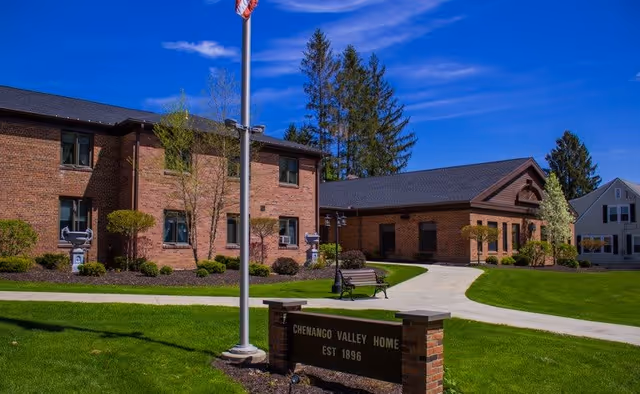 Exterior view of Chenango Valley Home, a brick building with two stories and a single-story wing, surrounded by well-maintained green lawns, trees, and shrubs under a clear blue sky. A flagpole with an American flag stands near a sign that reads 'Chenango Valley Home Est 1896'.