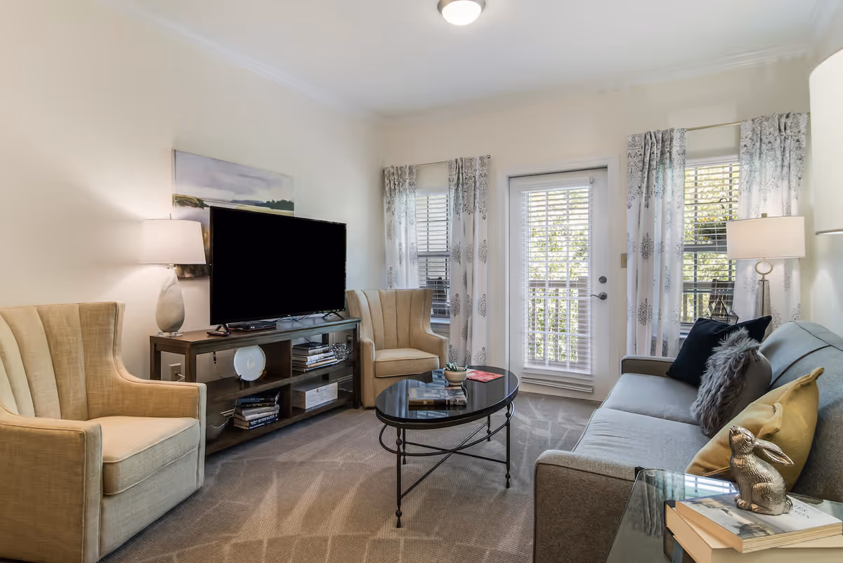 Well-lit living room with a sofa, two armchairs, TV on a console and a round coffee table in front of a glass door.