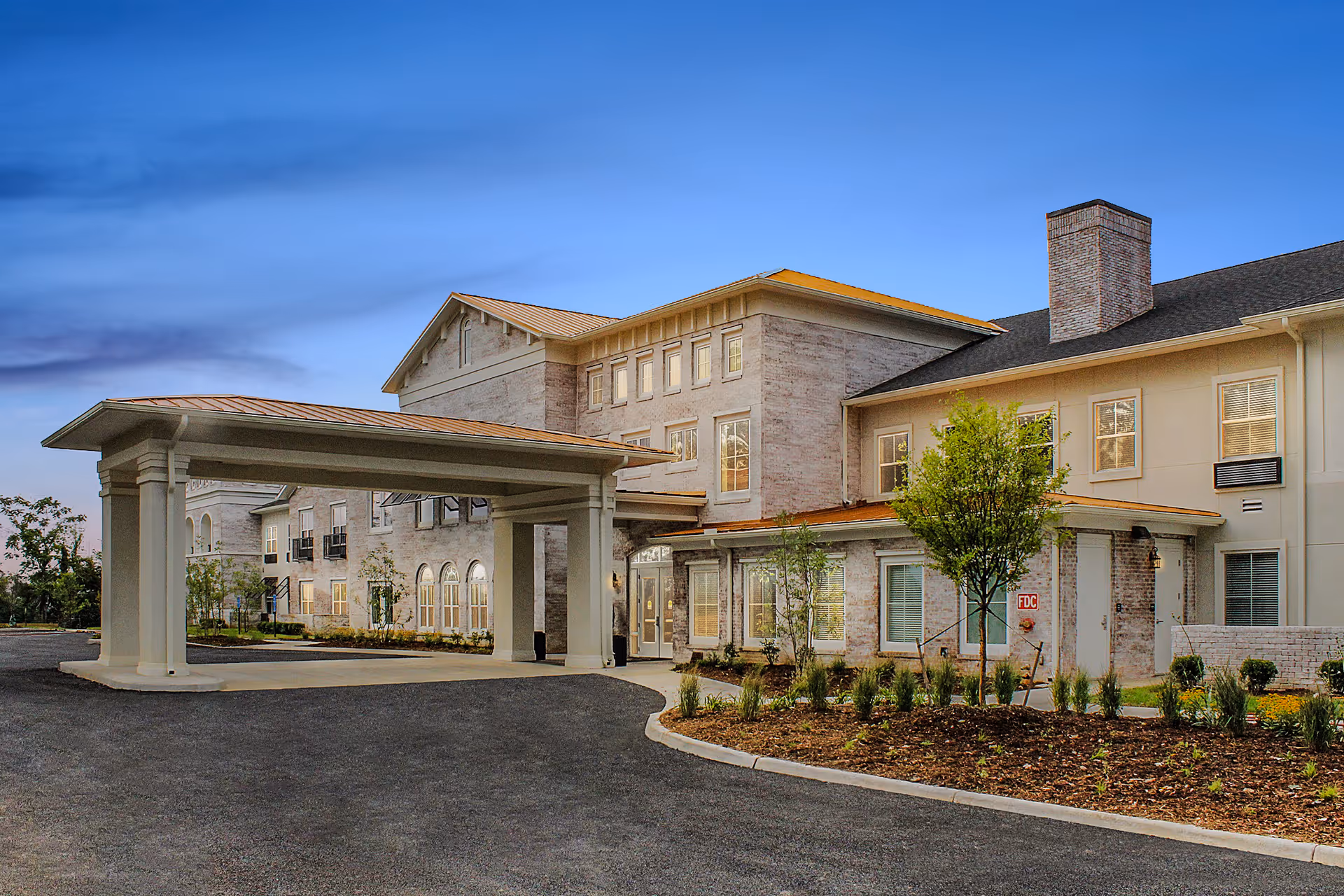 Exterior view of The Blake at Charlottesville senior living facility during dusk, showing a large covered entrance with columns, multiple windows, a brick chimney, and landscaped greenery along the driveway.