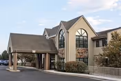 Exterior view of a two-story senior living facility building with a covered entrance, large arched windows, and surrounding shrubs under a partly cloudy sky.