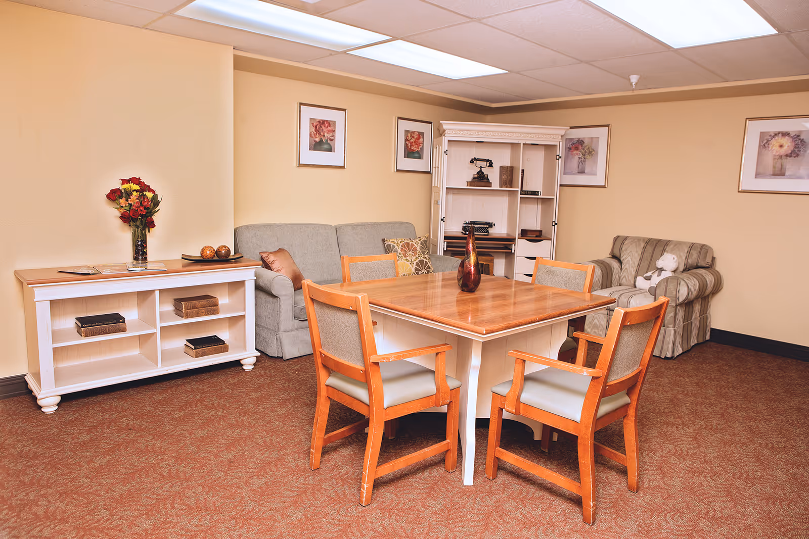 A furnished common room with a wooden table and chairs, a sofa and armchair, shelving, and framed artwork on the walls.
