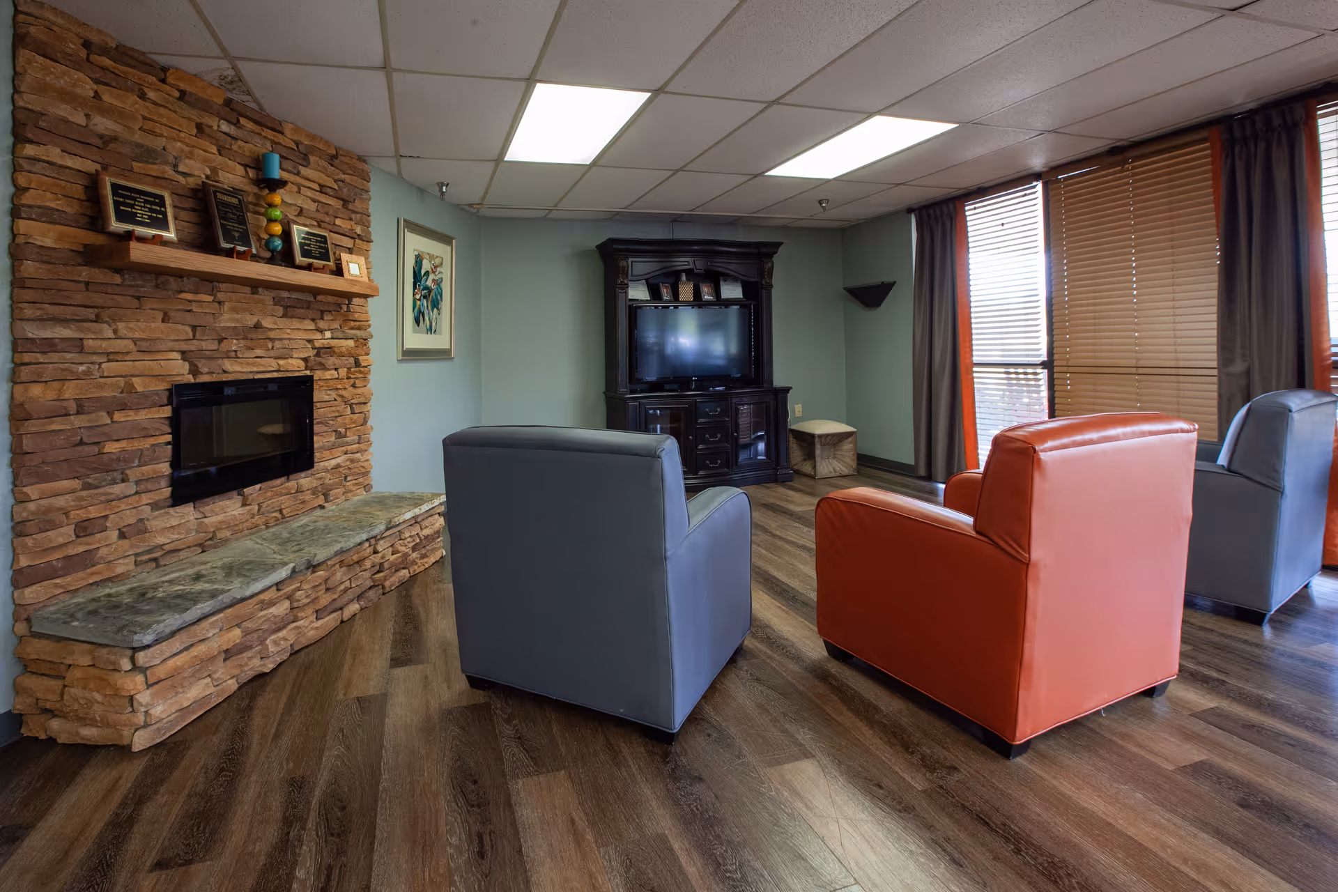 A cozy living room with a stone fireplace on the left wall, a wooden mantel with plaques and decorative items above it, and three armchairs in front of a TV cabinet. The room has wood flooring, light blue walls, and large windows with brown blinds and dark curtains on the right side.