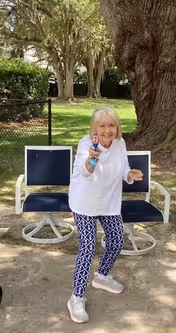 An elderly woman wearing a white long-sleeve shirt and patterned blue and white pants is standing outdoors on a sandy surface, holding a toy water gun and smiling. Behind her are two blue cushioned outdoor chairs with white frames, a large tree trunk, and green grass with trees in the background.
