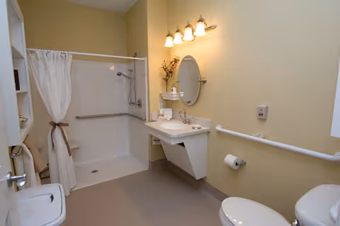 A senior-friendly bathroom featuring a walk-in shower with a white curtain and grab bars, a wall-mounted sink with a round mirror above it, a toilet with a grab bar beside it, and beige walls with overhead lighting.