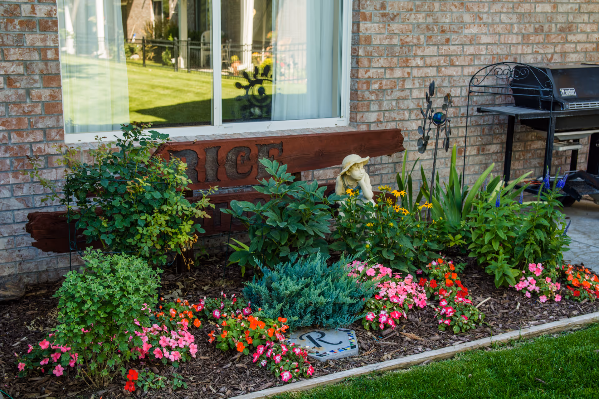 Colorful flower bed with a small garden statue and wooden sign in front of a brick building window and outdoor grill.