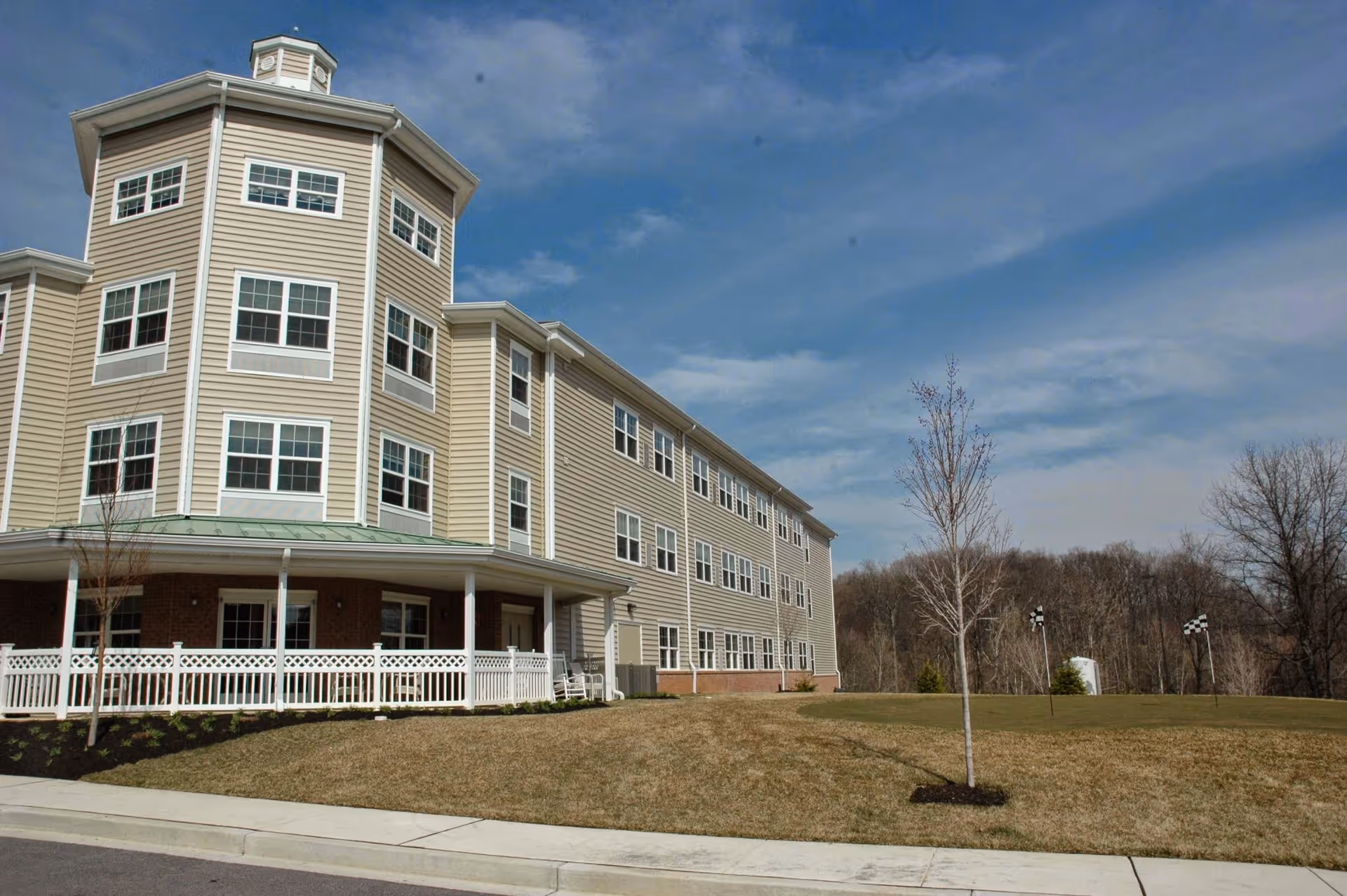 Exterior view of a multi-story senior living facility building with beige siding and white trim, a covered porch with white railing, a small lawn with young trees, and a partly cloudy blue sky.