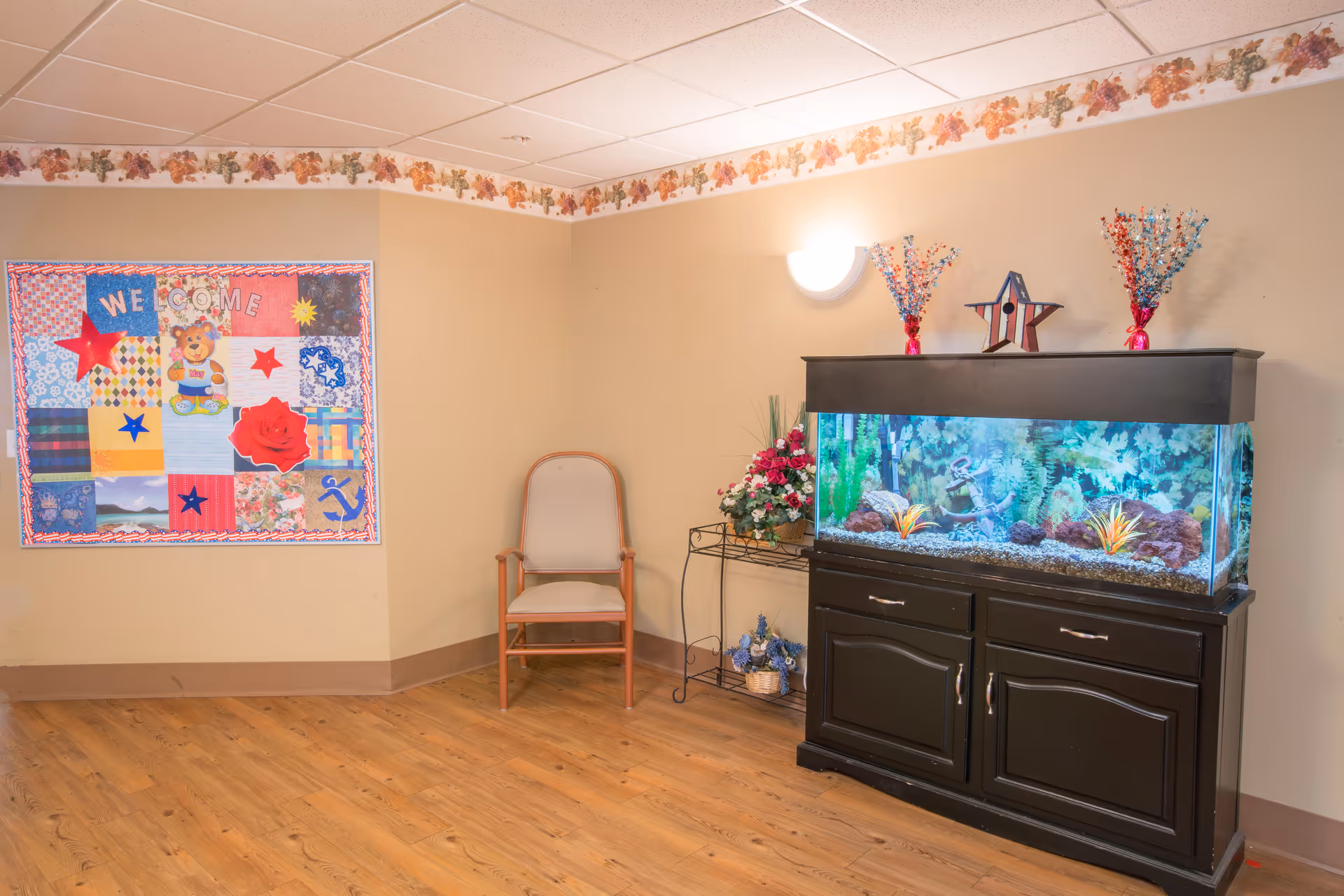 Interior corner of a room with a wooden floor and beige walls. A single wooden chair with a cushioned seat is placed against the wall. To the right, there is a large black aquarium filled with water, plants, and decorative items, sitting on a matching black cabinet. Above the aquarium are two vases with red, white, and blue decorations and a star ornament. On the left wall, there is a colorful quilted wall hanging with the word 'WELCOME' and various patterns and images. The ceiling has white tiles and a floral border runs along the top of the walls.