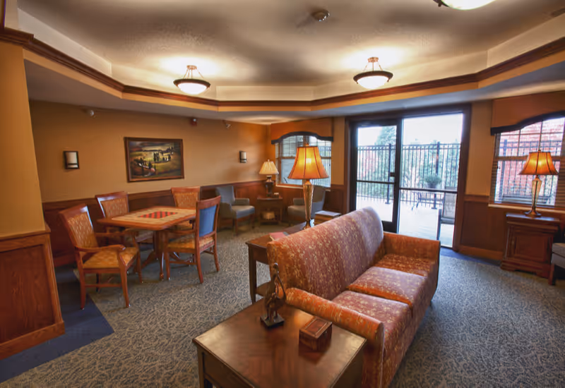 A cozy living room area in an assisted living facility featuring a patterned sofa, wooden side tables with lamps, a table with four chairs, additional seating near the windows, and large glass doors leading to an outdoor patio.
