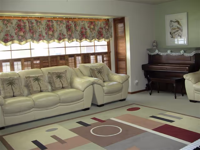 Living room with cream leather sofas and palm-printed pillows, a patterned area rug, a large window with a floral valance, and an upright piano.