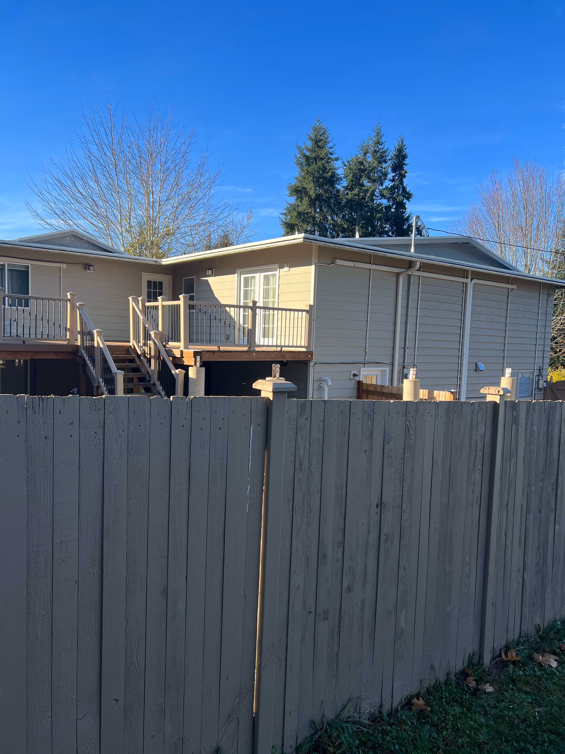 Exterior view of a beige two-story building with a wooden fence in the foreground. The building has a small balcony with railings and stairs leading down. Trees without leaves and evergreen trees are visible in the background under a clear blue sky.