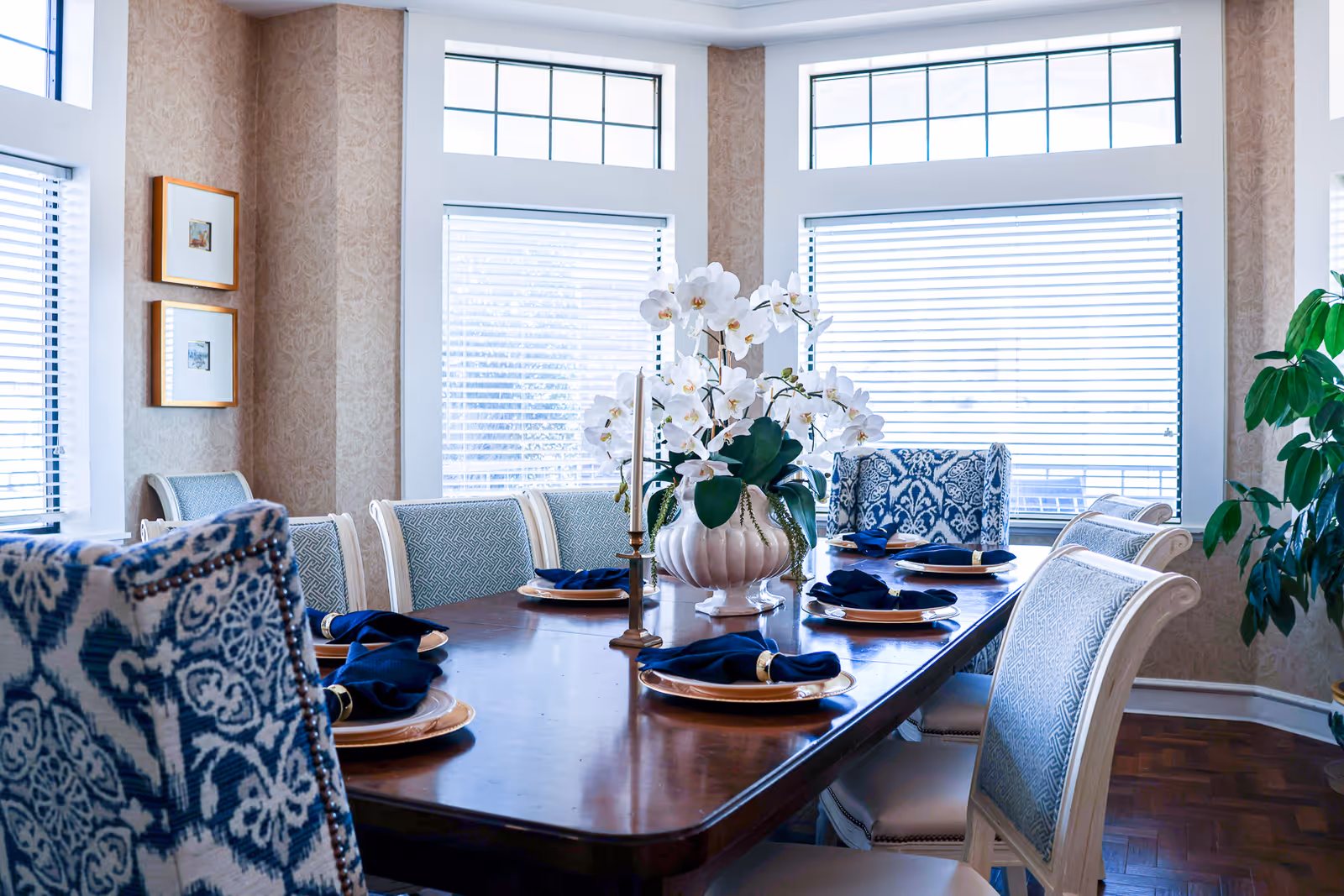 Sunlit dining room with a wooden table set with plates, navy napkins, a central orchid arrangement, and upholstered chairs by large windows.