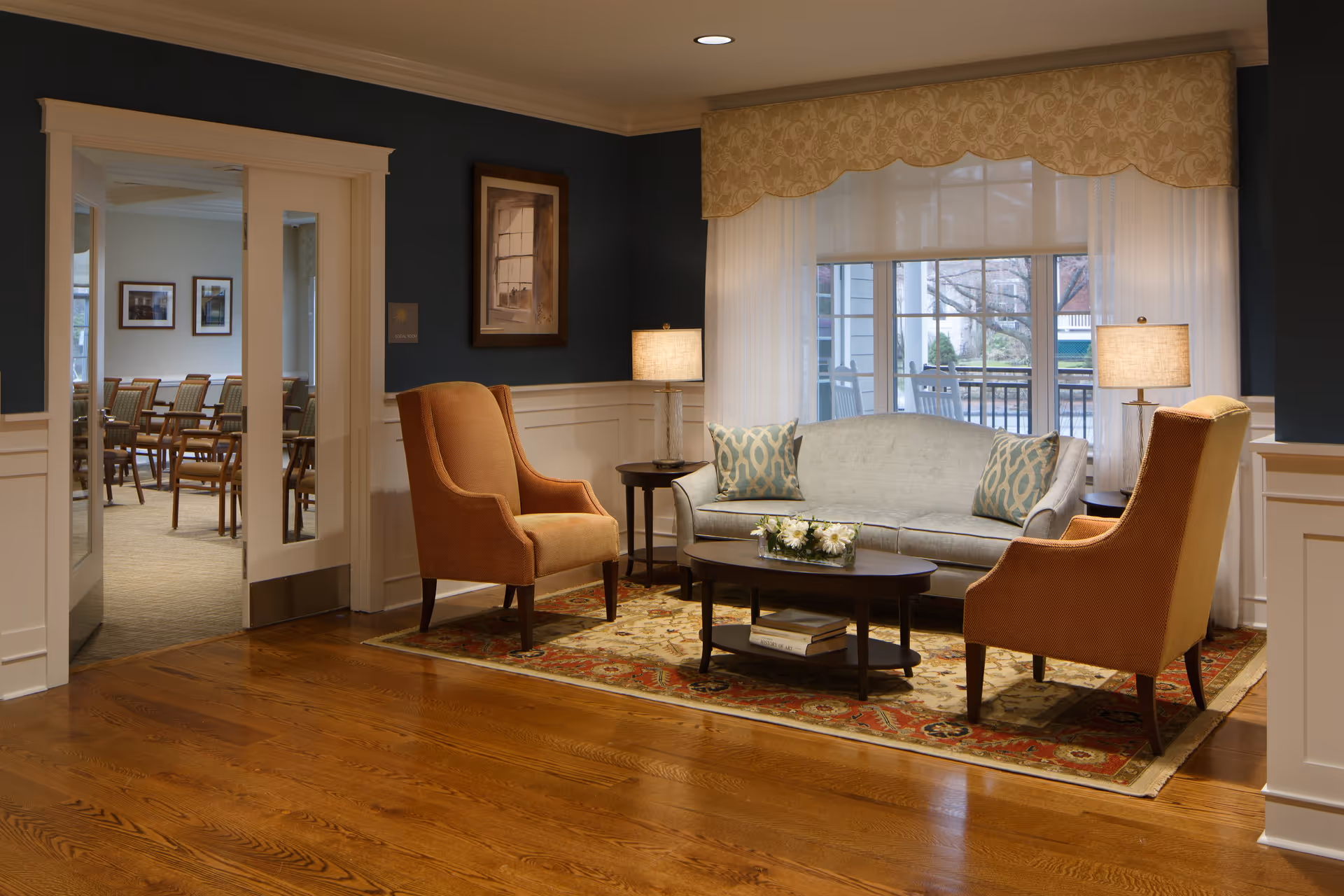 A cozy sitting area in a senior living facility with two orange armchairs and a light gray sofa with patterned pillows arranged around a dark wooden coffee table on a decorative rug. Two table lamps with beige shades flank the sofa. A large window with sheer white curtains and a patterned valance lets in natural light. In the background, an adjacent room with multiple wooden chairs is visible through open double doors.