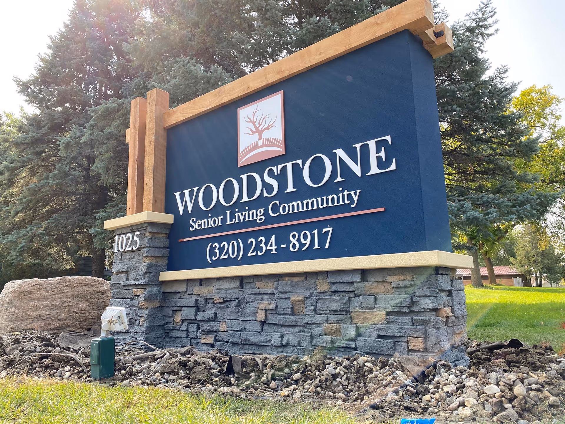 Outdoor sign for Woodstone Senior Living Community with a phone number (320) 234-8917, surrounded by trees and grass.