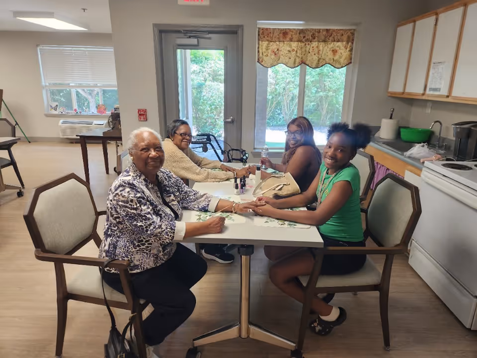 Four women sitting around a table in a room with a kitchen area, two of them are older adults and two are younger, engaging in a manicure activity with nail polish bottles on the table. The room has large windows with a view of greenery outside.