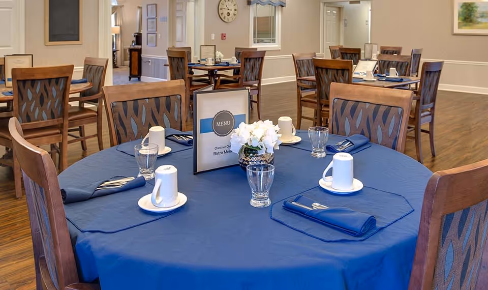 A dining room in a senior living facility with round tables covered in blue tablecloths. Each table is set with white coffee cups on saucers, glasses, blue cloth napkins with silverware, and a small floral centerpiece. A menu card is placed on the table in the foreground. Wooden chairs with patterned backs surround the tables. The room has wooden flooring and neutral-colored walls with some framed artwork and a clock.
