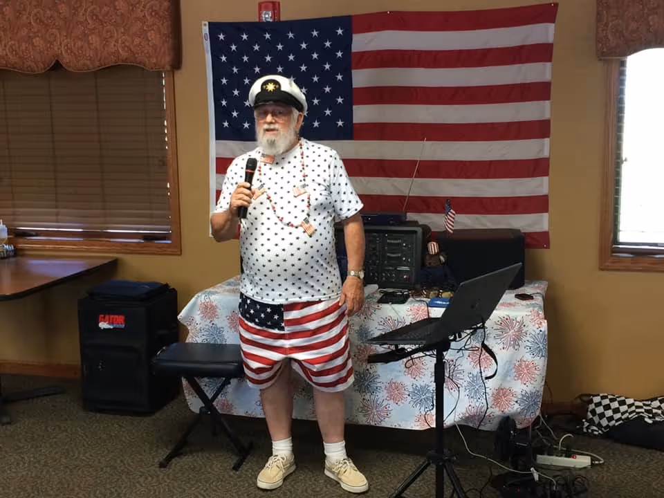 An elderly man wearing a captain's hat, a white shirt with blue stars, and American flag-themed shorts stands indoors holding a microphone. Behind him is a table covered with a festive tablecloth and an American flag hanging on the wall. There is audio equipment and a laptop on a stand nearby. The room has beige walls, windows with blinds, and carpeted flooring.