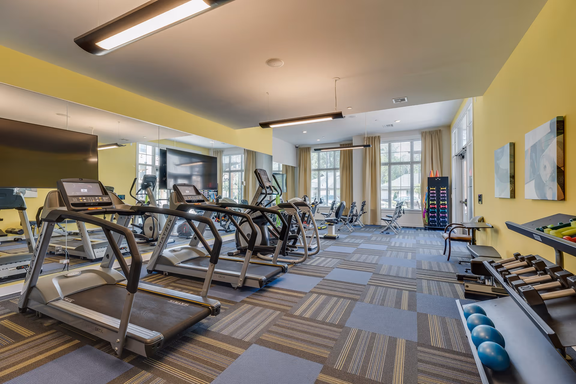 A spacious fitness room with several treadmills, exercise bikes, and weight racks. The room has large windows with beige curtains, yellow walls, and a patterned carpet floor. Mirrors cover one wall, and there are various exercise equipment and weights neatly arranged.