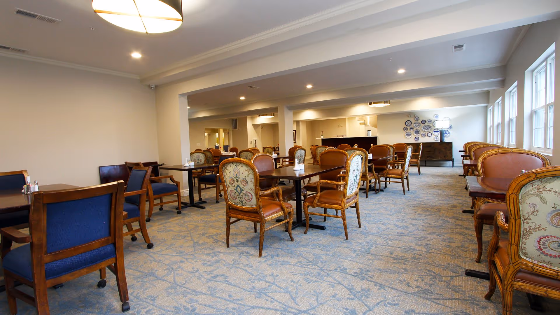 A spacious dining room in a senior living facility with multiple wooden tables and chairs arranged neatly. The chairs have a mix of blue upholstery and patterned backs with wooden frames. The room is well-lit with ceiling lights and natural light coming through large windows on the right side. The carpet has a subtle floral pattern, and there is a sideboard with decorative plates on the far wall.