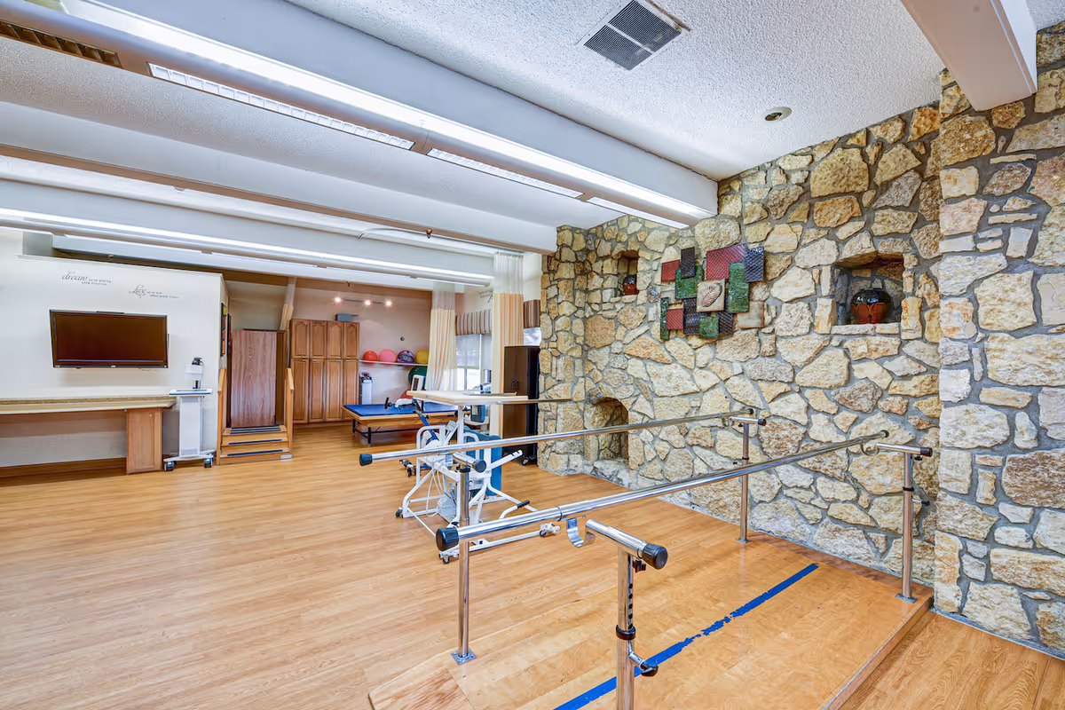 A spacious room with wooden flooring and a stone wall decorated with small alcoves and colorful art pieces. The room contains parallel bars for physical therapy or rehabilitation exercises, a treatment table, and a wall-mounted TV. The ceiling has fluorescent lighting, and there are cabinets and exercise balls in the background.