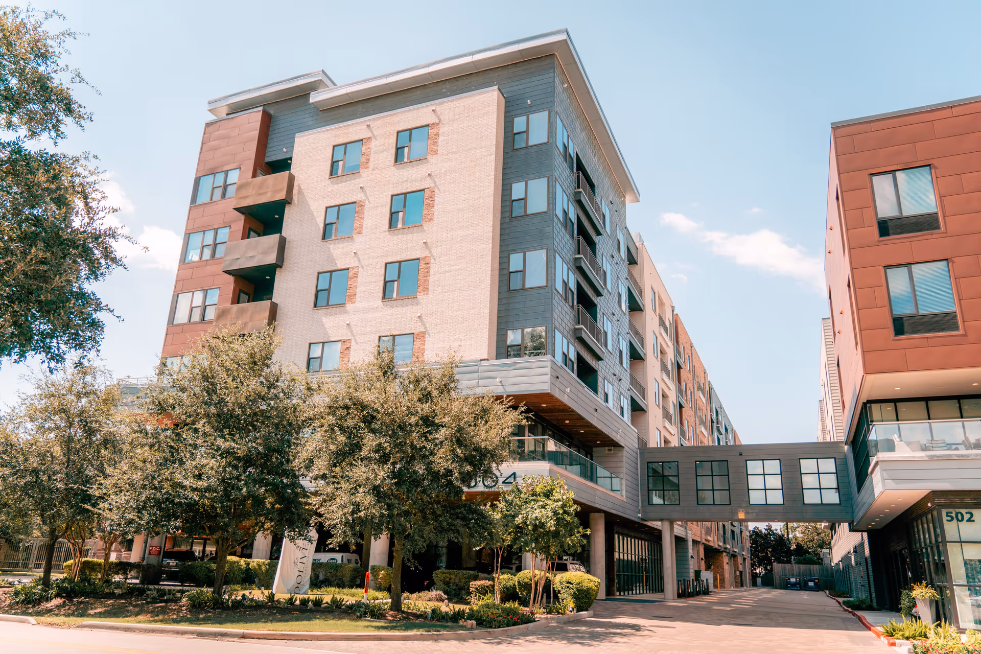 Exterior view of a modern multi-story residential building with a sky bridge connecting two sections. The building features large windows, balconies, and a landscaped area with trees and shrubs in front. The sky is clear and blue.