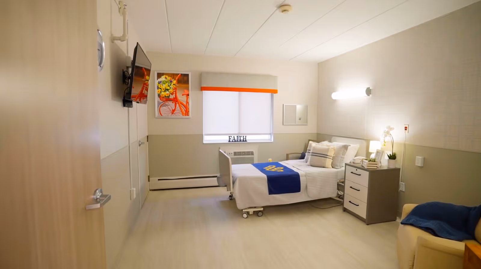 Accessible rehab patient room with a single bed, bedside table, wall-mounted TV, window, and armchair.