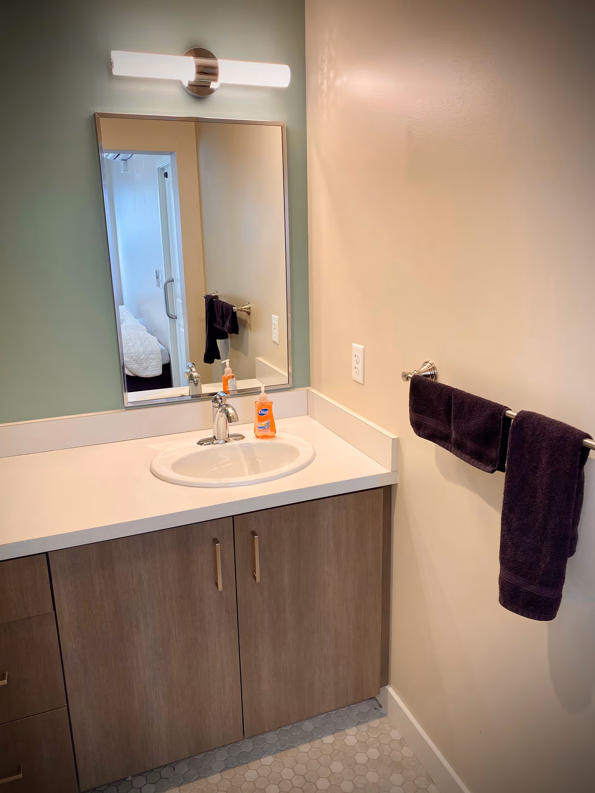 A bathroom vanity with a sink, mirror, overhead light, soap dispenser on the counter and a dark towel hanging on a wall rack.
