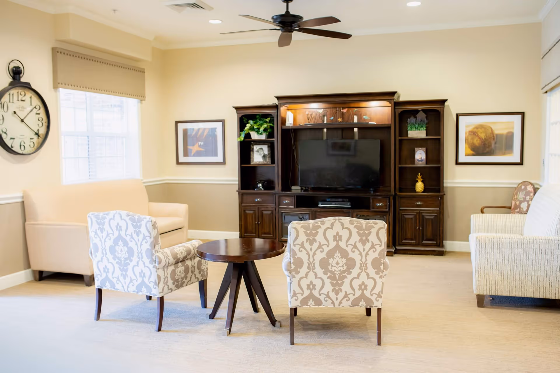 Bright common living room with patterned armchairs around a round table facing a large wooden entertainment center and television.