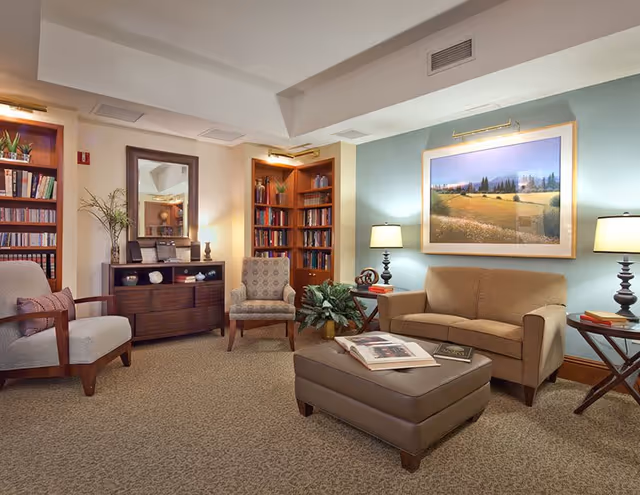 A cozy living room area with a beige sofa, two armchairs, a large ottoman with books on it, two side tables with lamps, a wooden cabinet with a mirror above it, and built-in bookshelves filled with books and decorative items. A large framed landscape painting hangs on the wall, and there is a potted plant near the sofa.