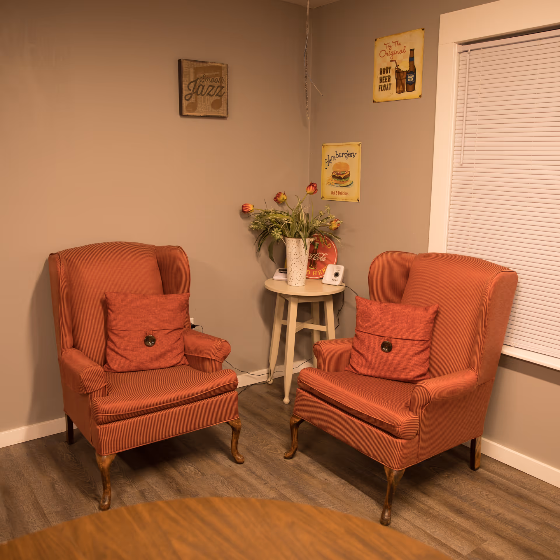 Two red upholstered armchairs with matching cushions are positioned around a small round side table with a vase of flowers and a small device on it. The room has beige walls with three vintage-style signs hanging, and a window with closed blinds is visible on the right side.