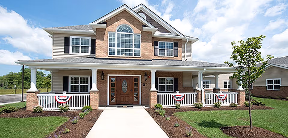 Front exterior view of a two-story senior living facility building with a large porch, white railings decorated with red, white, and blue bunting, and a well-maintained lawn with small plants and a young tree.