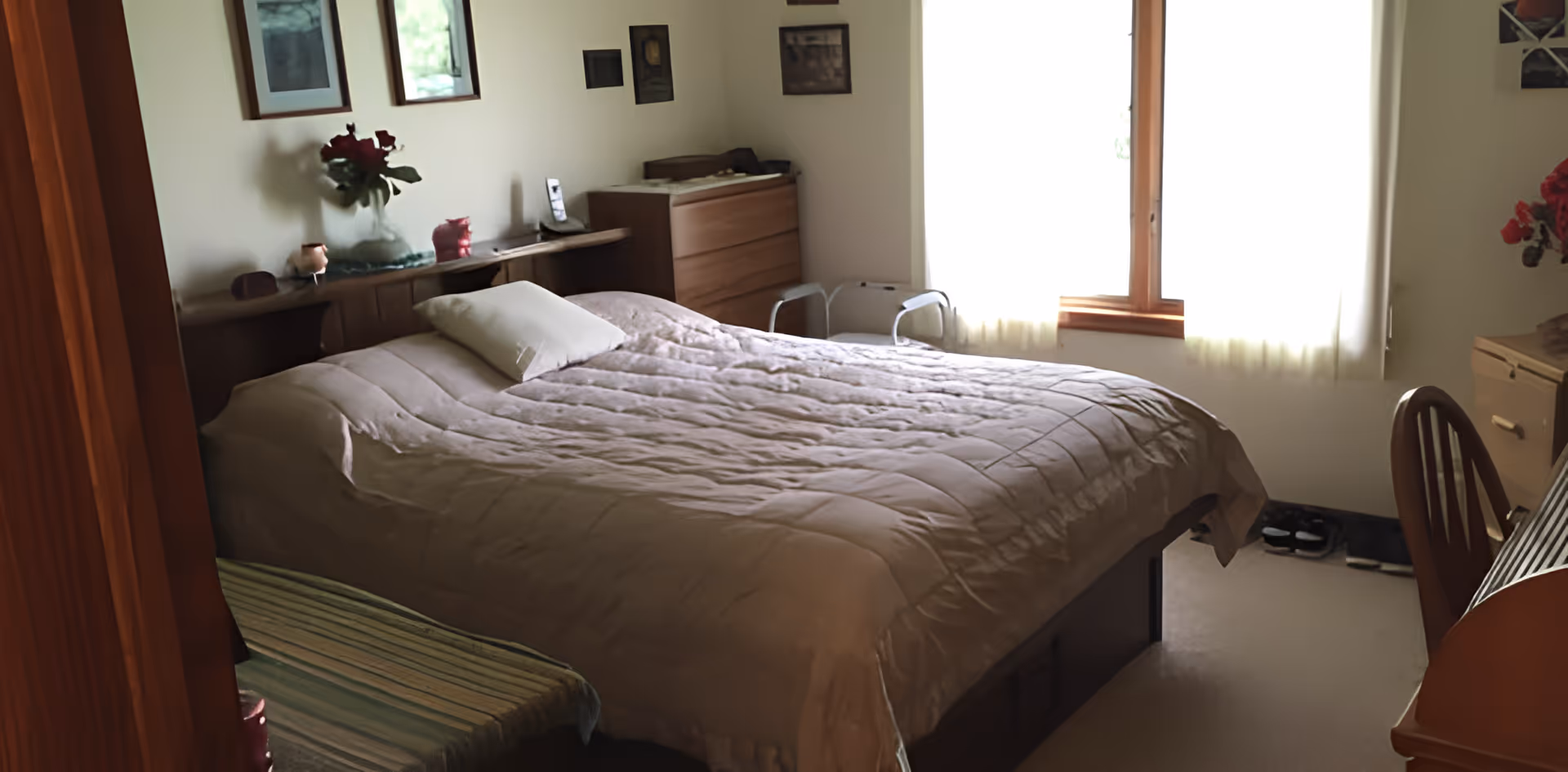 A cozy bedroom with a large bed covered in a beige quilt and a white pillow. The room has a wooden dresser, a chair, a small bench with a striped cushion, and framed pictures on the walls. Light filters in through a window with sheer white curtains.