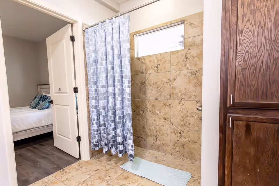 View of a bathroom with a tiled walk-in shower featuring a blue and white patterned shower curtain and a light blue bath mat on the floor. To the left, an open door reveals a bedroom with a bed that has white bedding and decorative pillows. On the right side, there is a wooden cabinet with silver handles.