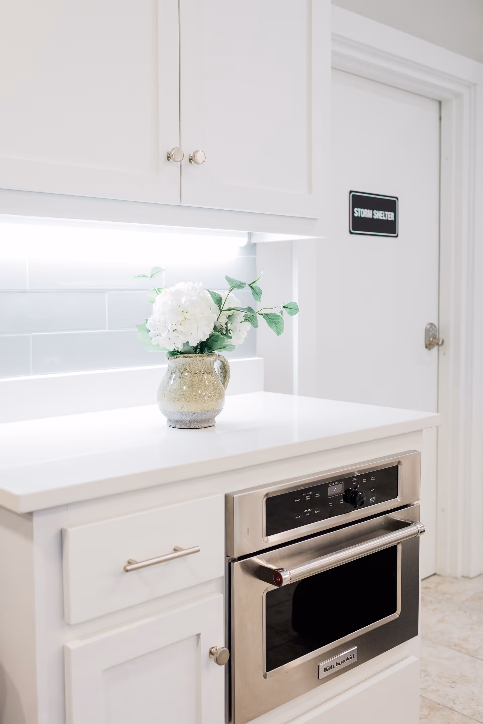 A modern kitchen countertop with a built-in stainless steel KitchenAid oven. On the white countertop, there is a ceramic vase holding white flowers and green leaves. Above the counter, white cabinets are visible, and in the background, there is a white door with a black sign that reads 'STORM SHELTER'.