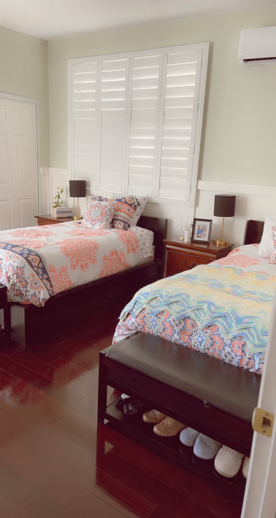 Bright bedroom with two twin beds, nightstands, white plantation shutters, and a bench with shoes under it.
