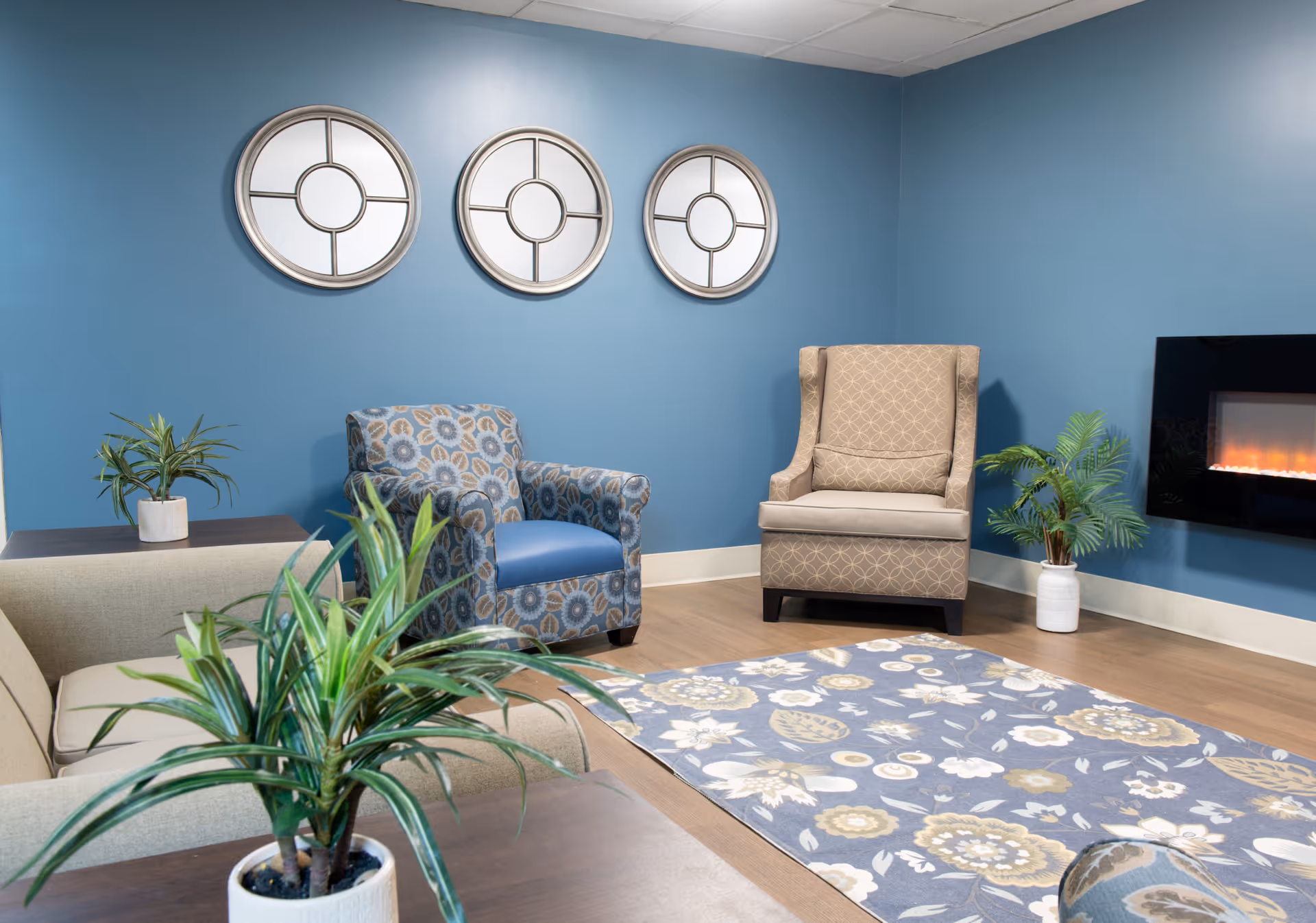 A cozy living room with blue walls featuring three round decorative mirrors. The room has a beige sofa, a patterned blue armchair, a beige wingback chair, a floral area rug, two potted plants, and a modern electric fireplace mounted on the wall.