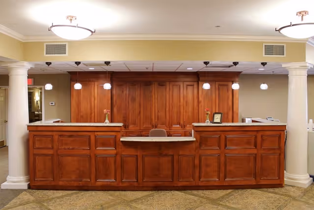 Reception desk area with a large wooden counter featuring multiple panels and a granite countertop. Behind the desk are wooden cabinets and a chair. The ceiling has recessed lighting and hanging pendant lights. Two white columns flank the desk on either side, and the floor is carpeted with a patterned design.