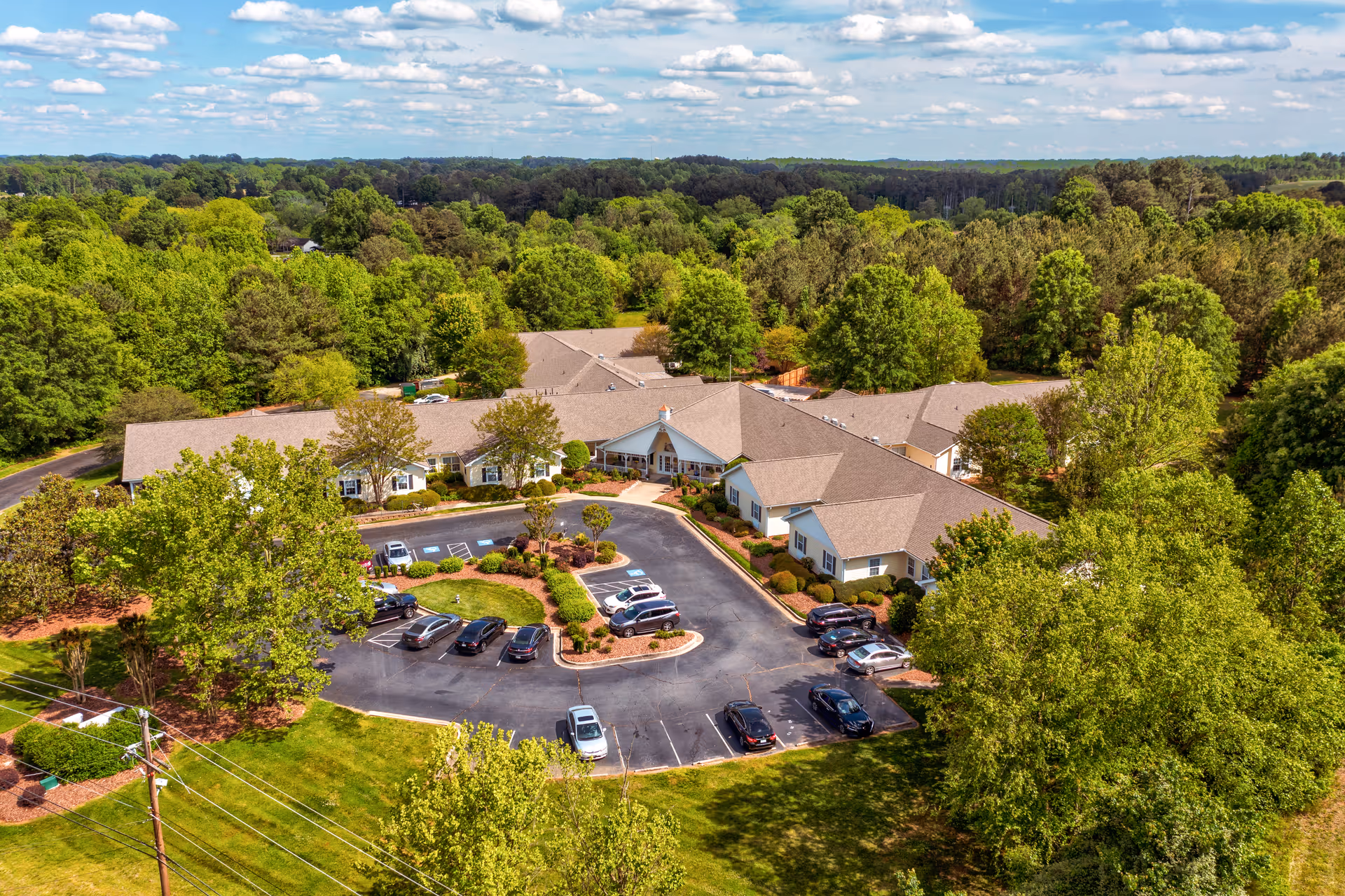 Aerial view of TerraBella Salisbury senior living facility surrounded by lush green trees and landscaping, with a parking lot in front containing several parked cars under a partly cloudy sky.