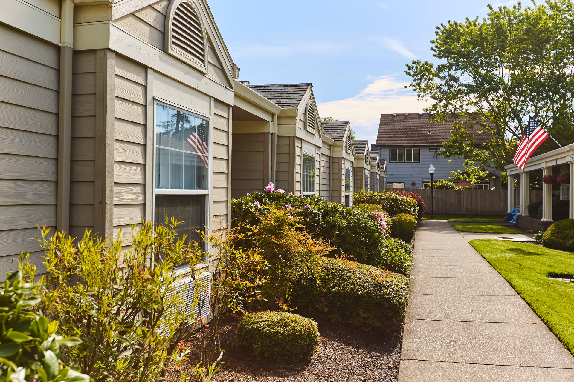 Exterior view of a senior living facility with beige siding, multiple windows, and well-maintained landscaping including bushes and flowers along a paved walkway. Two American flags are visible, one reflected in a window and one mounted on a porch. A large tree and a neighboring house are in the background under a partly cloudy sky.