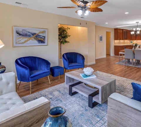 Bright open living room with blue velvet chairs, a wooden coffee table, ceiling fan, and a view into the dining area and kitchen.
