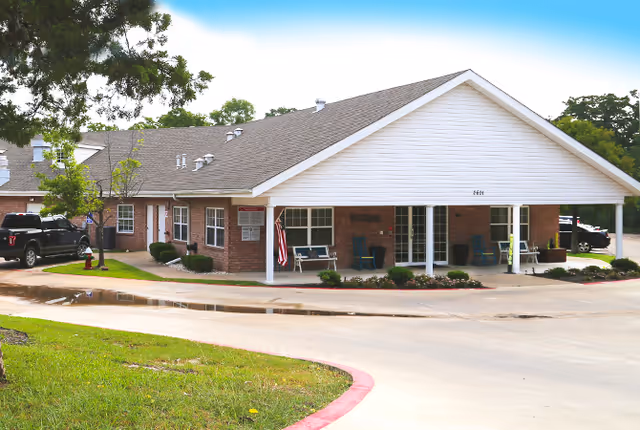 Exterior view of a single-story brick building with a large covered entrance, several windows, and a few parked cars nearby. The building is surrounded by a well-maintained lawn and trees under a clear blue sky.