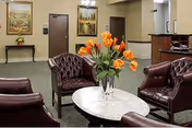 A cozy seating area in a care center with four dark brown leather armchairs arranged around a white marble oval coffee table holding a vase of orange tulips. The room has green carpet, beige walls, two closed brown doors, a painting on the wall, and a small table with a flower arrangement in the background.