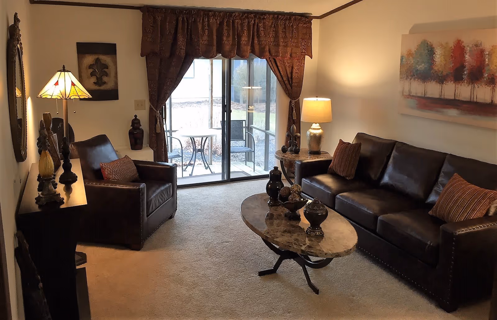 A cozy living room with a dark leather sofa and matching armchair, both adorned with decorative pillows. A marble coffee table with decorative items sits in the center on a beige carpet. There is a side table with a lamp next to the sofa and a console table with a lamp and decorative pieces against the wall. A large window with patterned curtains opens to a small outdoor patio with a table and two chairs. A colorful painting of trees hangs on the wall above the sofa.