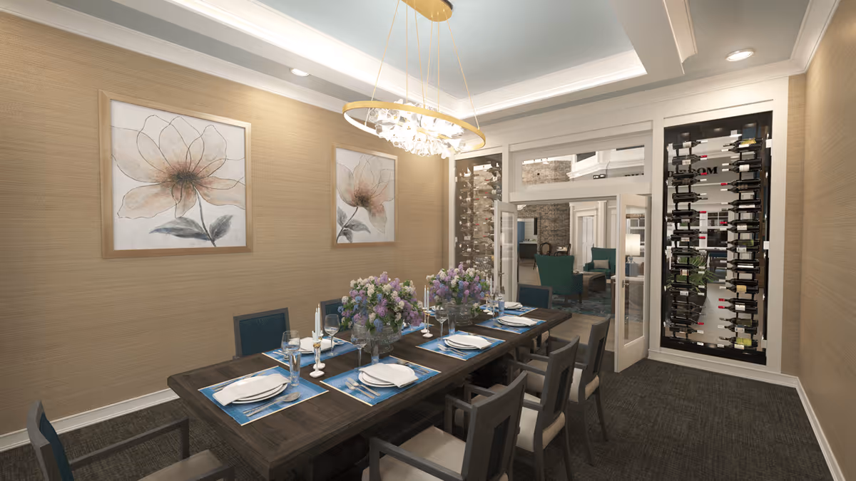 A formal dining room with a long dark wooden table set for eight people with white plates, silverware, wine glasses, and blue placemats. The table is decorated with three large floral centerpieces and white candles. The walls are beige with two large floral paintings. A modern chandelier hangs above the table. There is a glass wine rack filled with bottles on one wall and a doorway leading to a lounge area with green chairs.