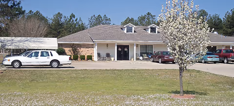 Exterior view of a single-story assisted living facility with parked cars and a blooming tree.
