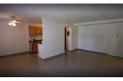 Empty living area with linoleum floor, a small kitchenette, ceiling fan, and an entry door.